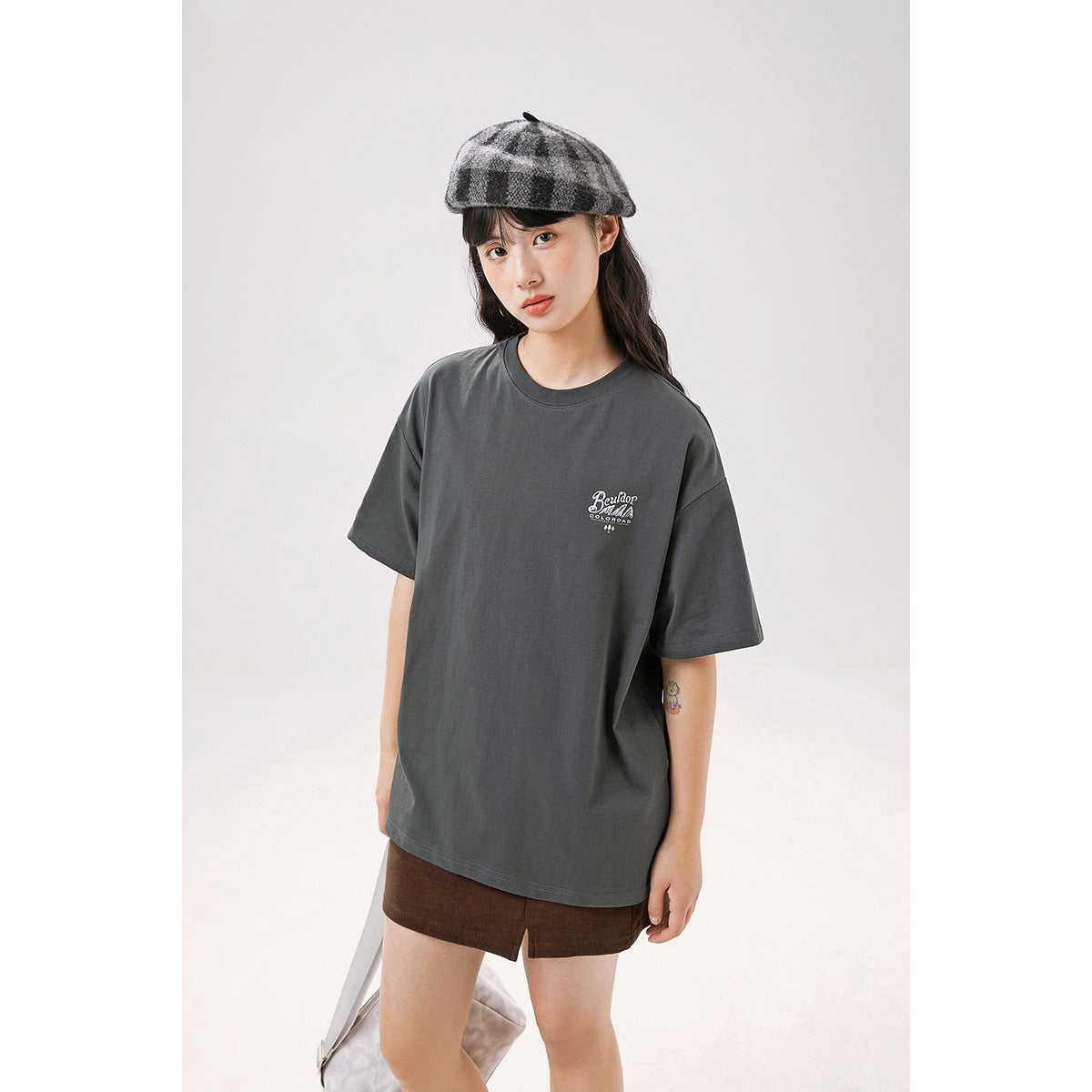 Model wearing an oversized charcoal gray T-shirt with a small white chest graphic, brown shorts, and a plaid cap.