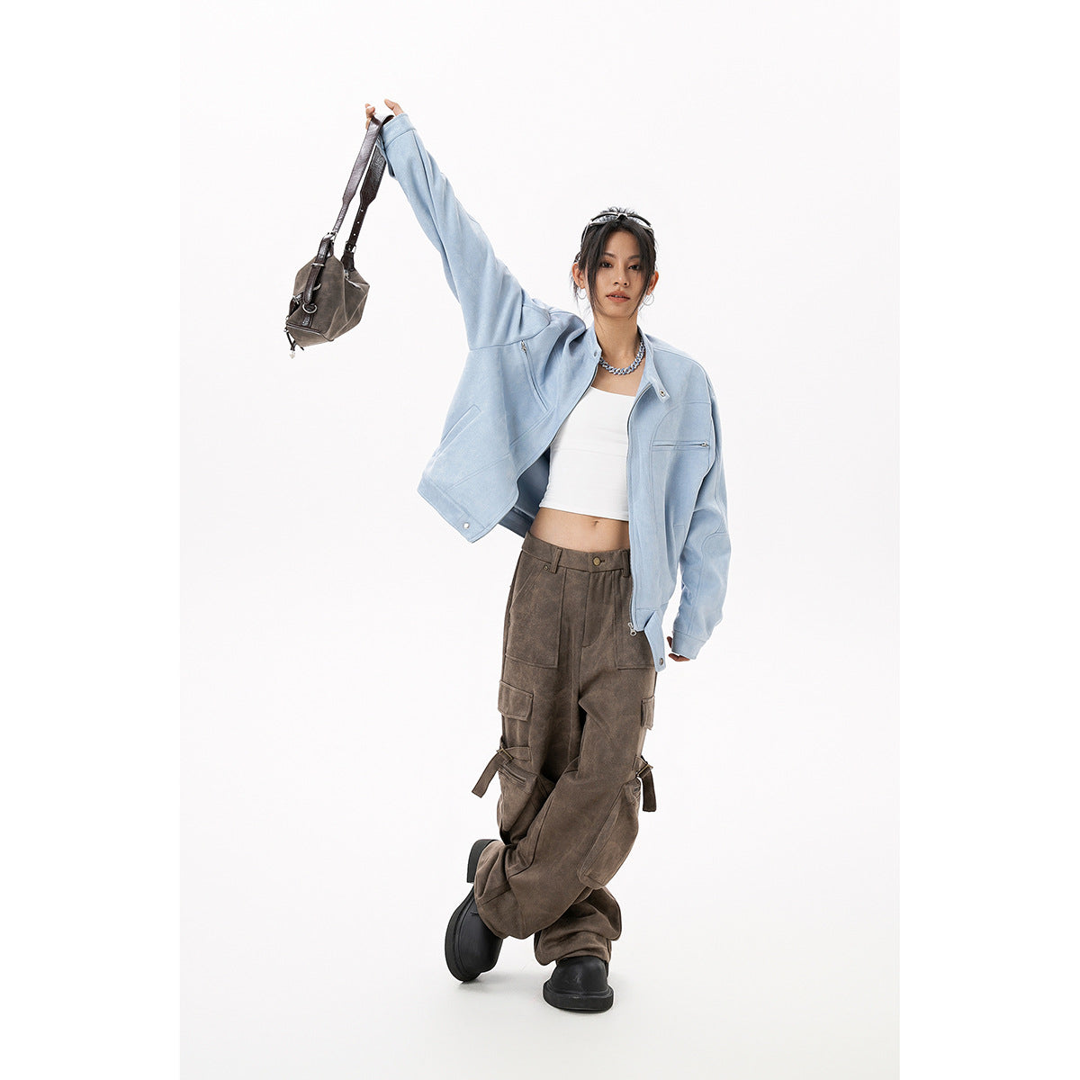 Model wearing an open light blue jacket and brown cargo pants, raising one arm while holding a brown bag.