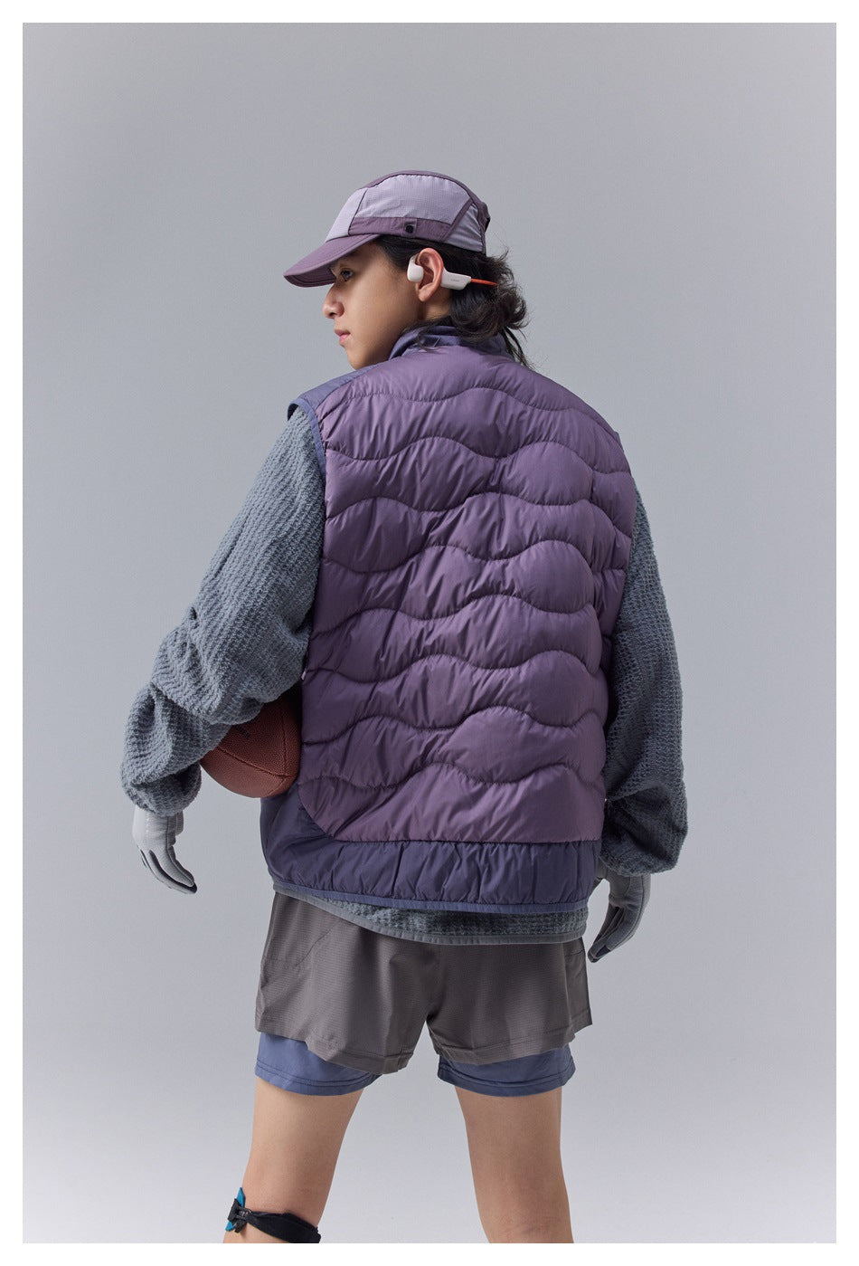 Model wearing a purple quilted vest with wavy stitching, shown from the back over a gray pullover.