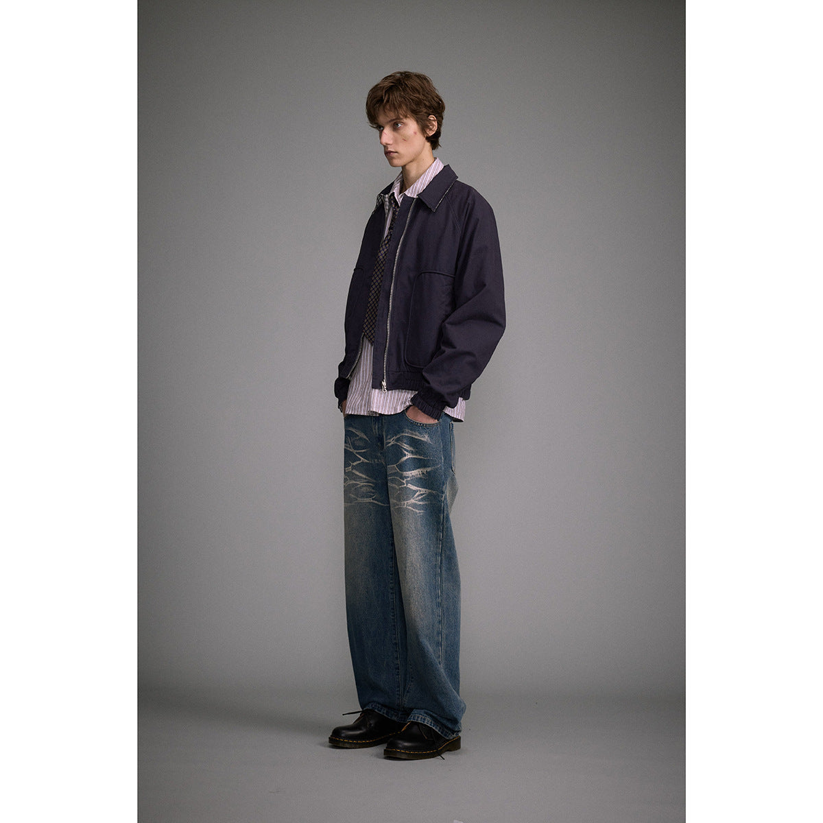 Model in a navy jacket, striped shirt and tie, wearing loose blue jeans with pale thigh designs.