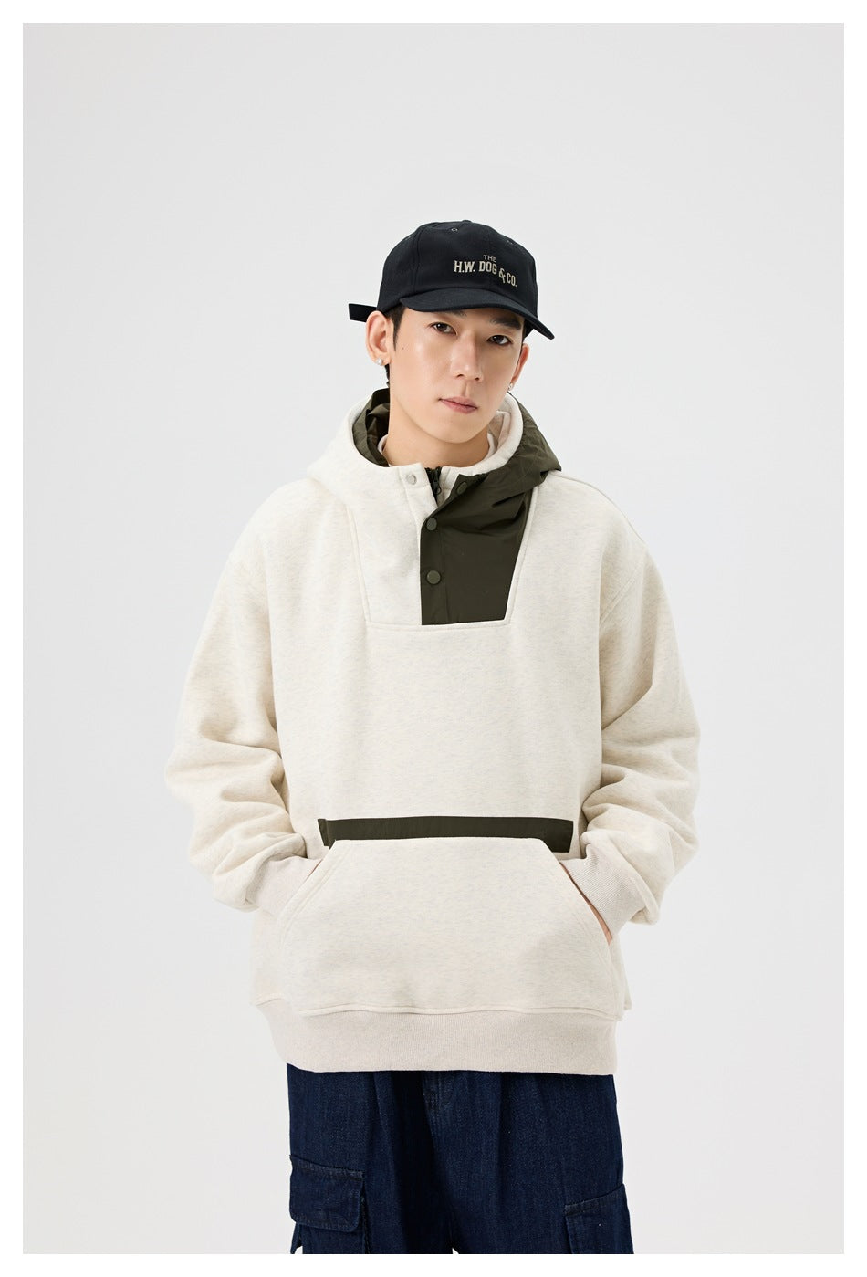 Model wearing oatmeal hooded sweatshirt with olive snap placket and pocket strip, black cap and dark blue cargo jeans