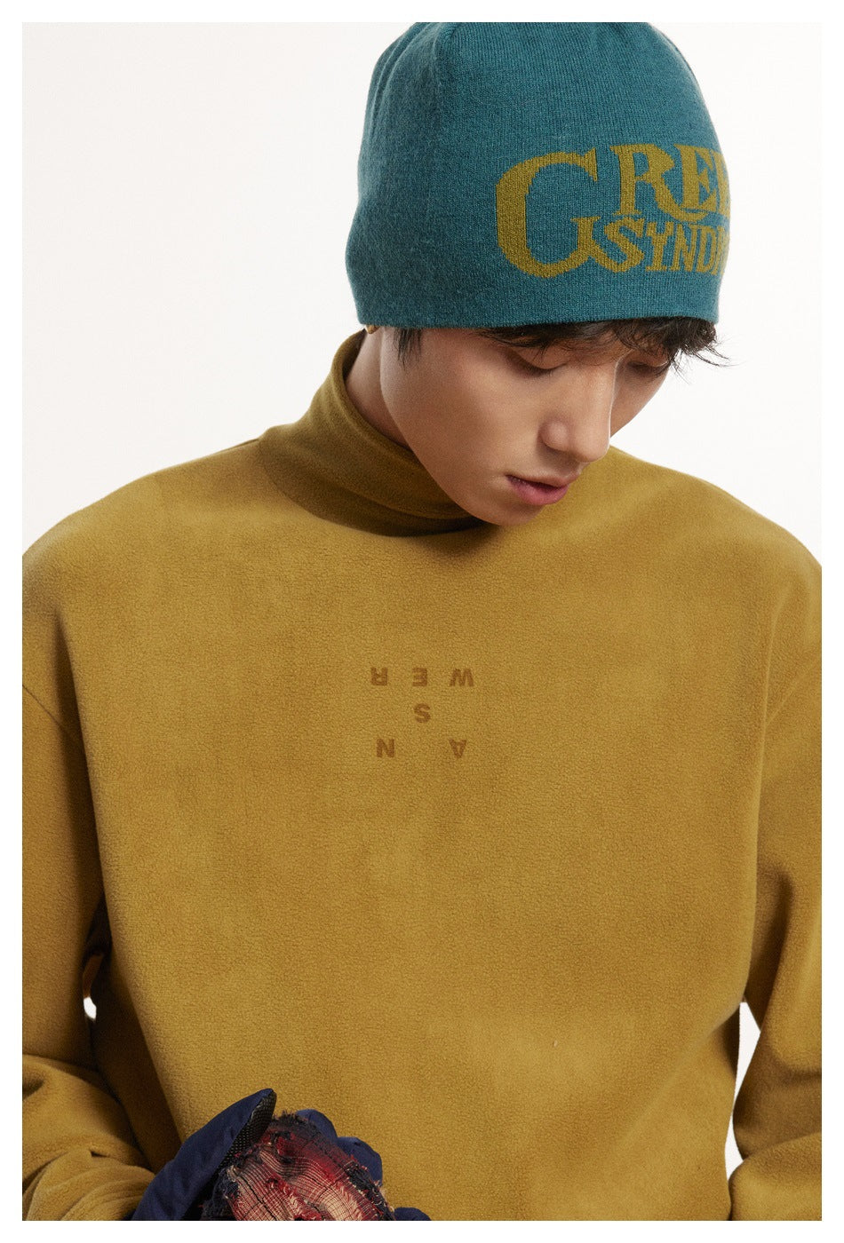 Close-up of a model wearing a mustard high-neck long-sleeve top with small chest lettering and a teal graphic beanie.