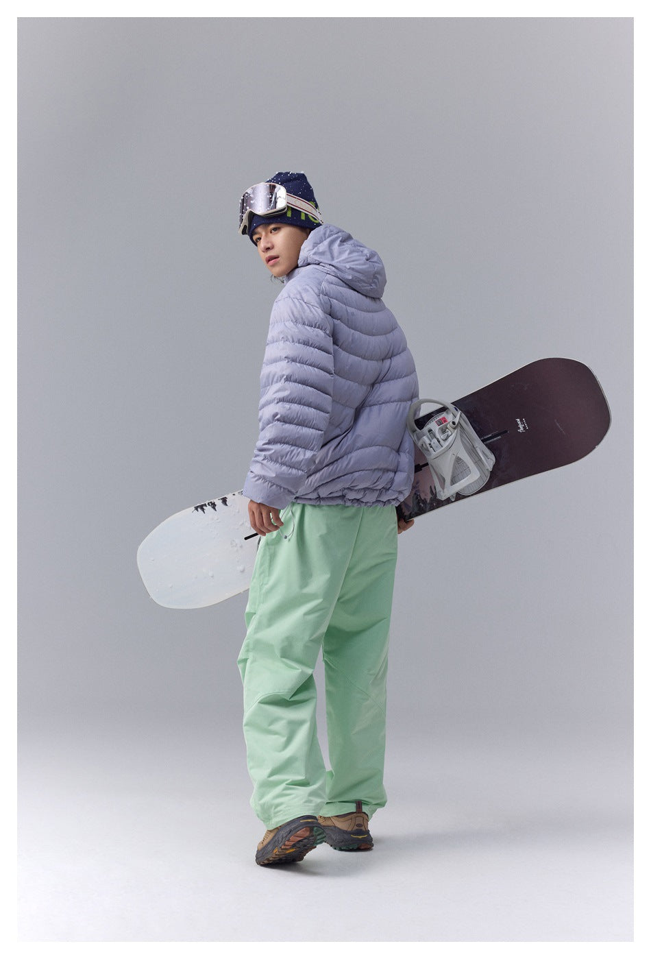 Model showing the back of a lavender hooded puffer jacket while carrying a snowboard.
