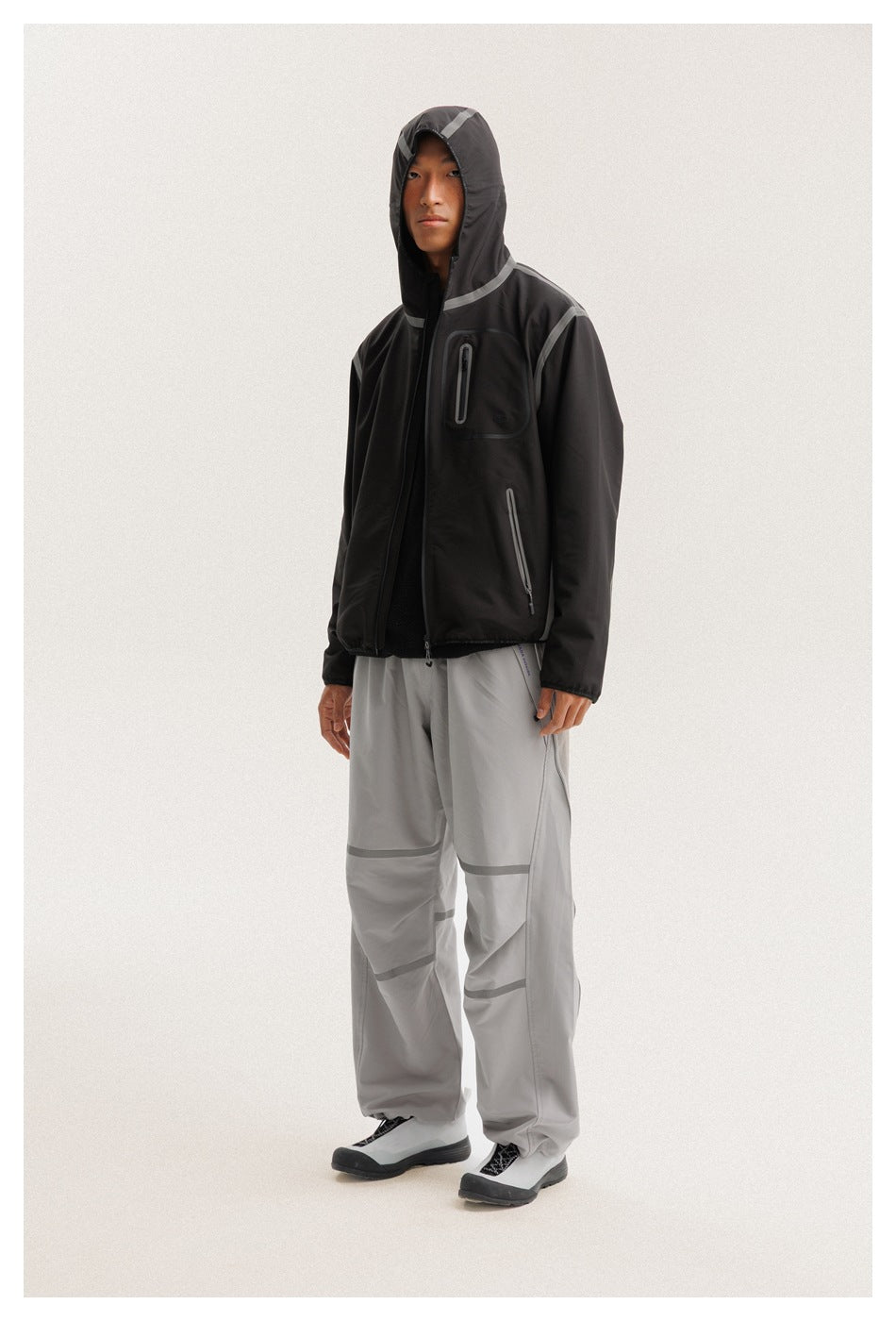 Full-length model wearing a black hooded jacket with gray trim and zip pockets, and loose light gray trousers.