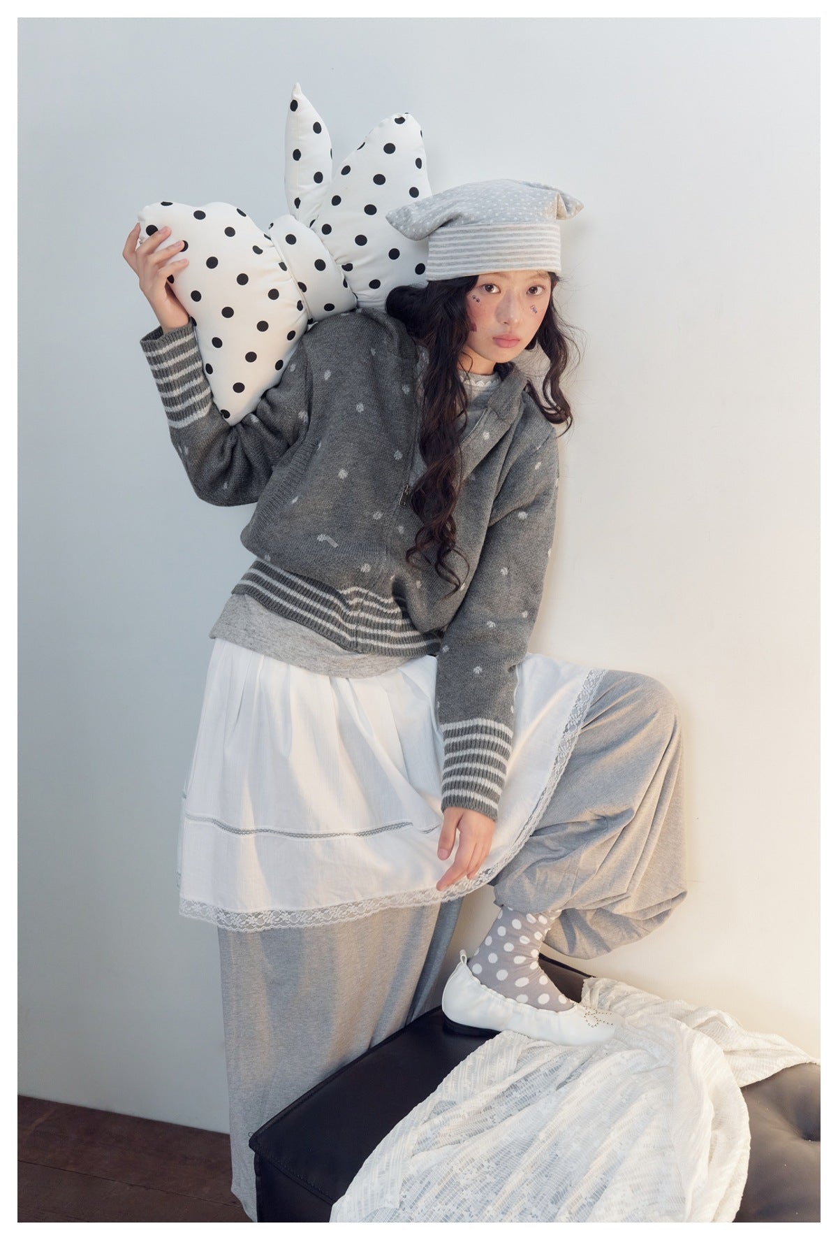 Model in a gray patterned zip cardigan and white tiered dress poses with one knee raised.