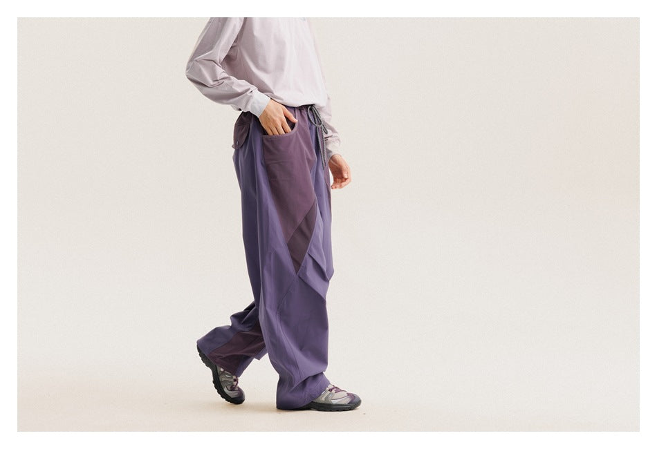 Side view of a model wearing loose purple paneled pants with contrasting geometric panels and a drawstring waist.