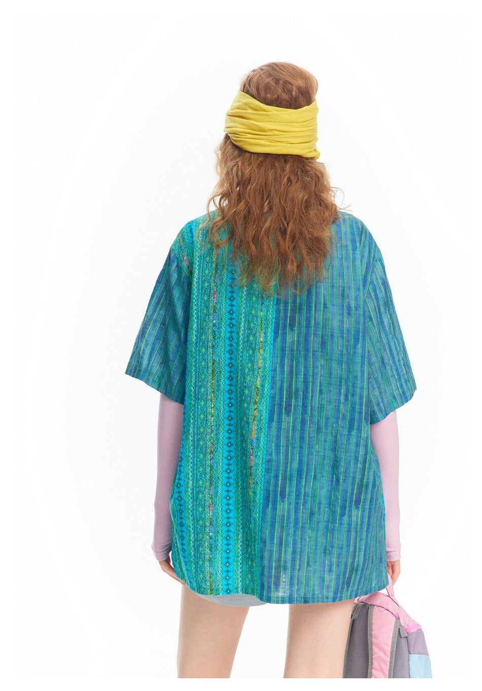 Rear view of a model wearing a loose blue-green patterned short-sleeve shirt with vertical stripes and decorative panels.
