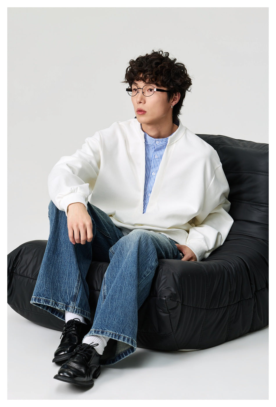 Man seated on a black chair wearing the white layered sweatshirt with striped shirt insert and blue jeans