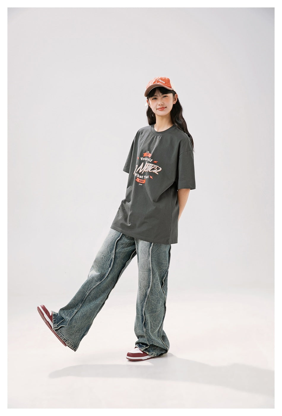 Person wearing a dark gray graphic T-shirt with an orange cap and wide-leg washed jeans, standing with one leg raised