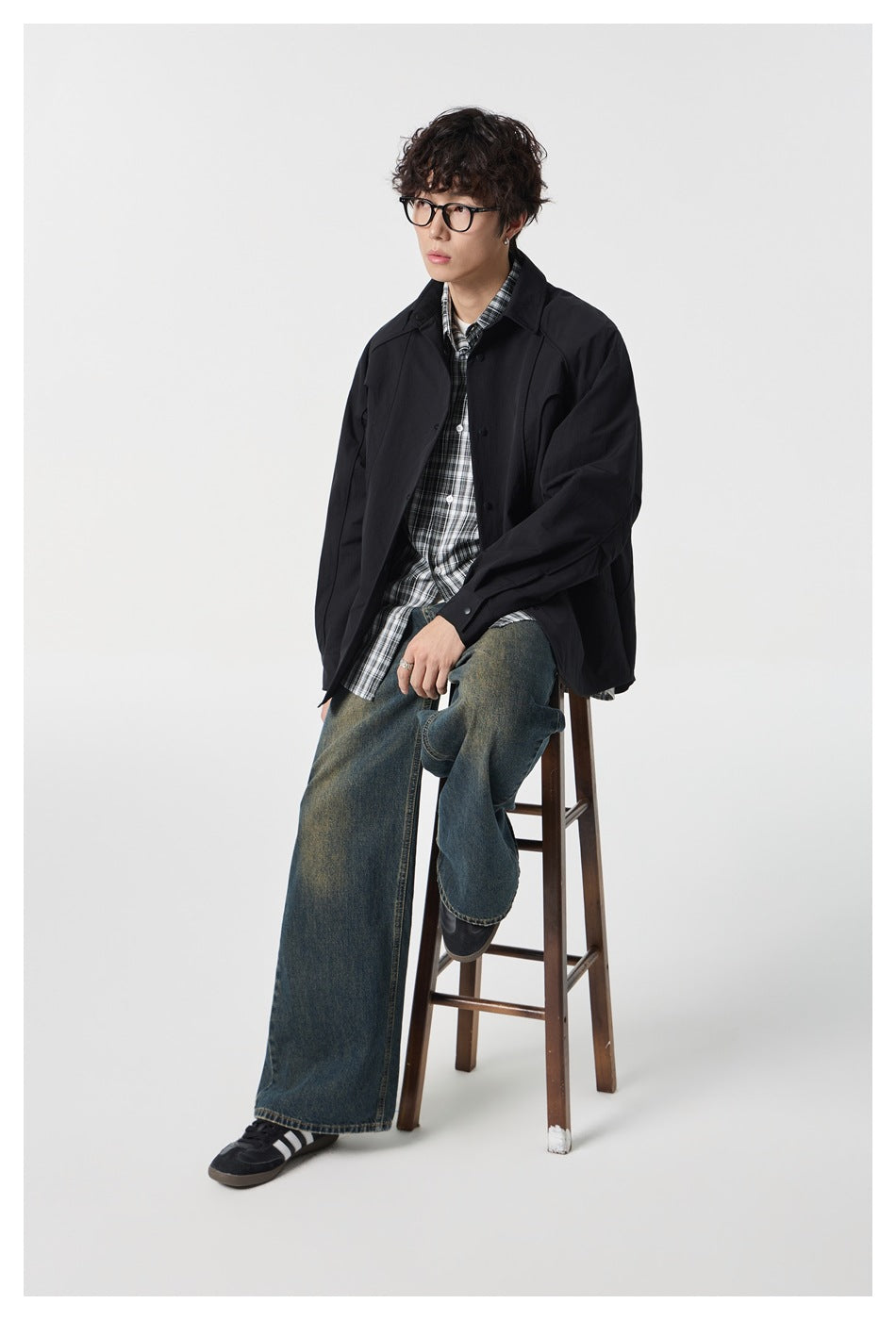 Model seated on a wooden stool wearing black shirt jacket over plaid shirt and wide-leg jeans