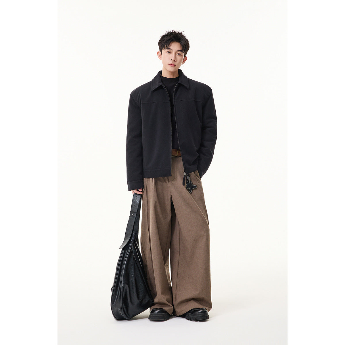 Full-length model wearing a black collared zip jacket with wide taupe trousers and carrying a black bag.