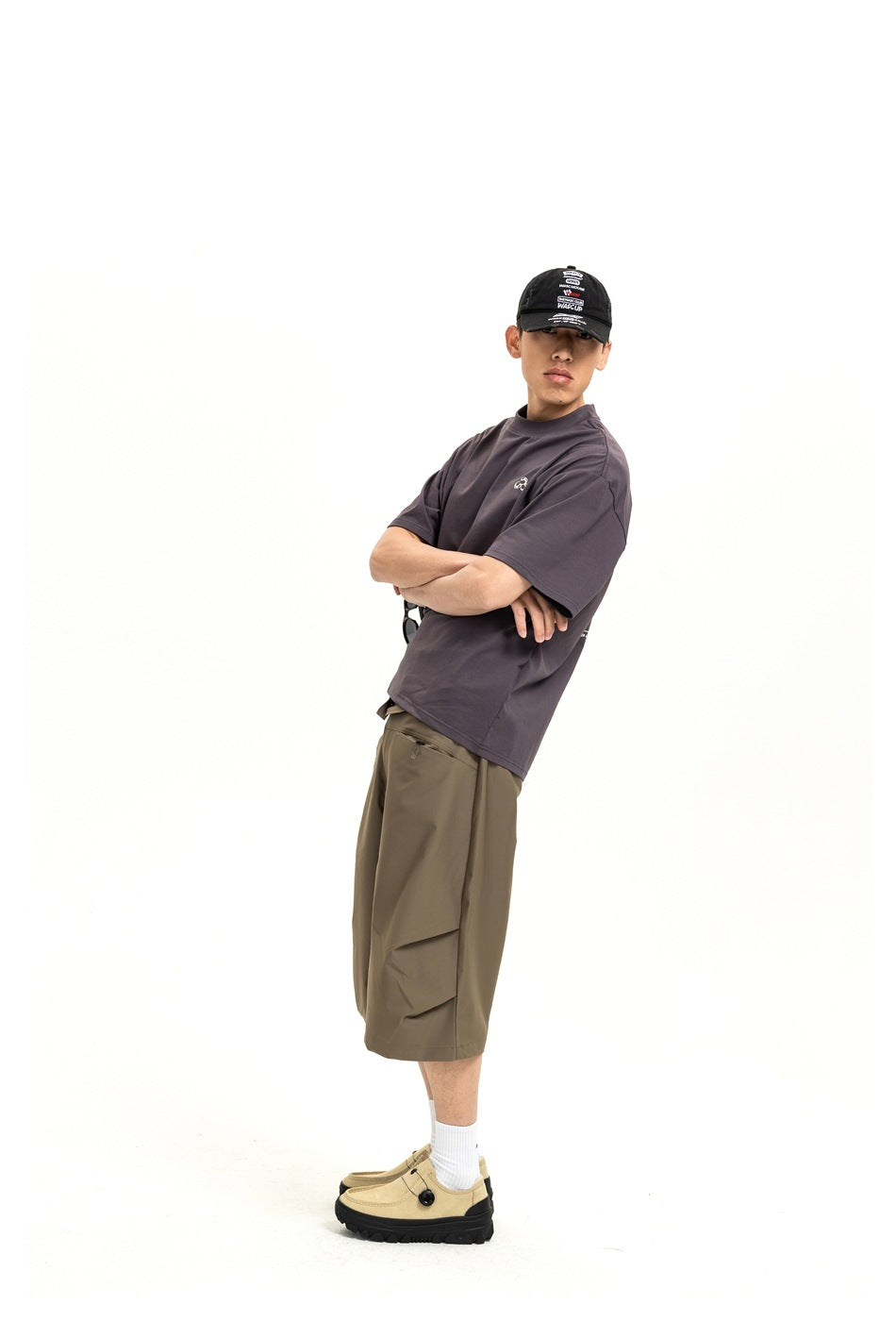 Side-view model wearing a charcoal T-shirt, olive-brown knee-length shorts, white socks and beige shoes.