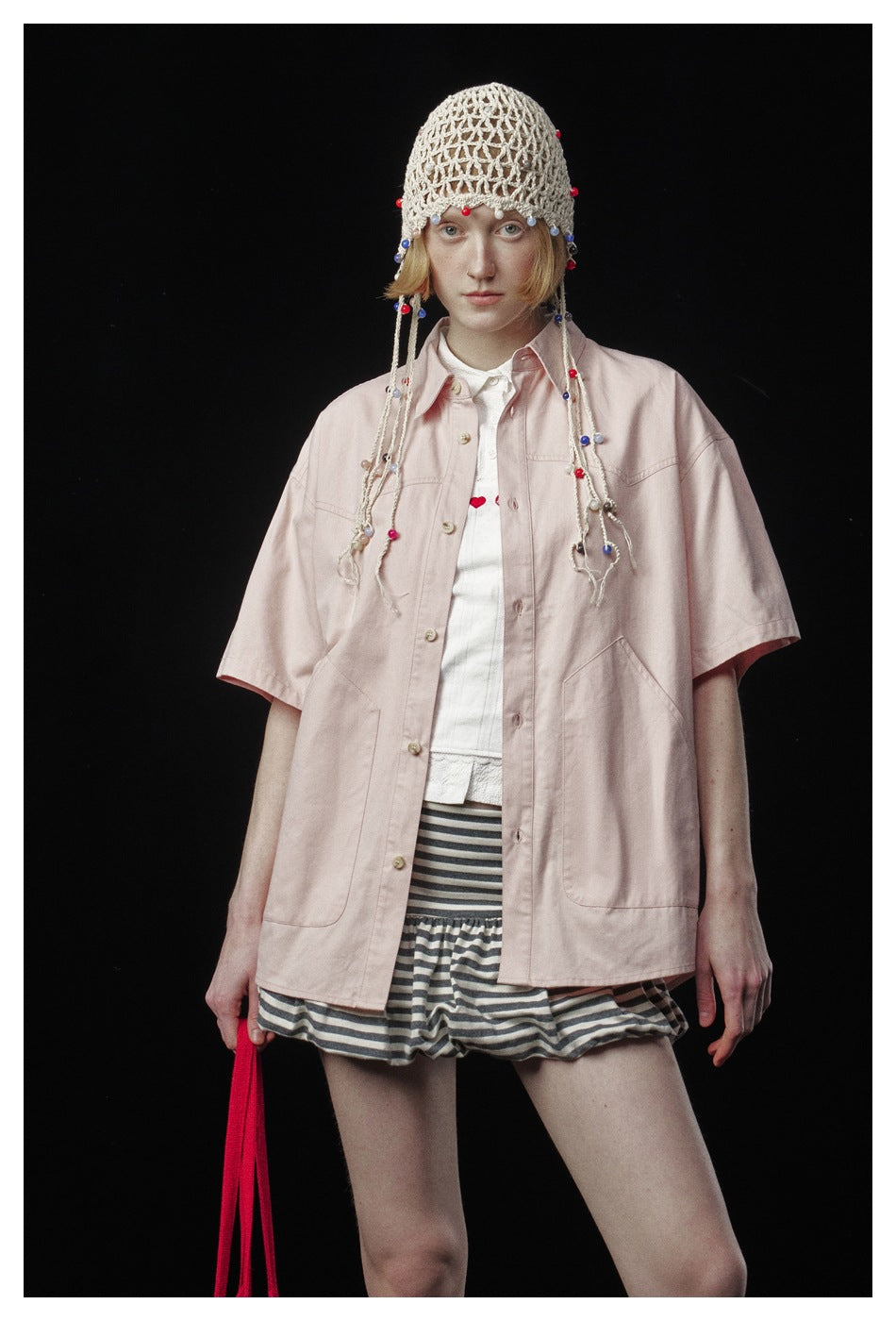 Close-up of a model in the pink short-sleeve shirt worn open over a white top with striped shorts and a beaded crochet cap