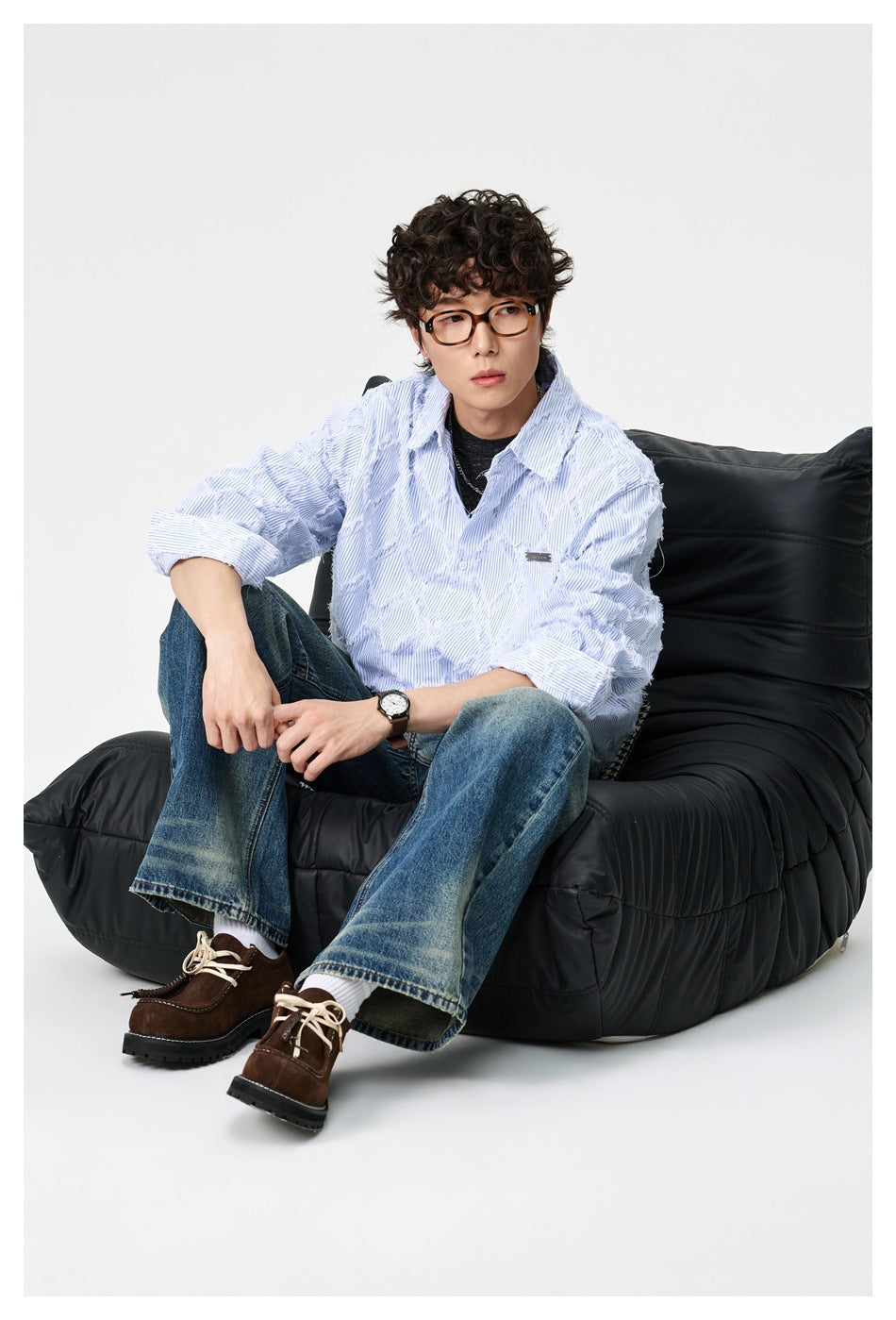 Model seated on a black lounge chair wearing a light blue pinstripe frayed-diamond shirt with wide-leg jeans