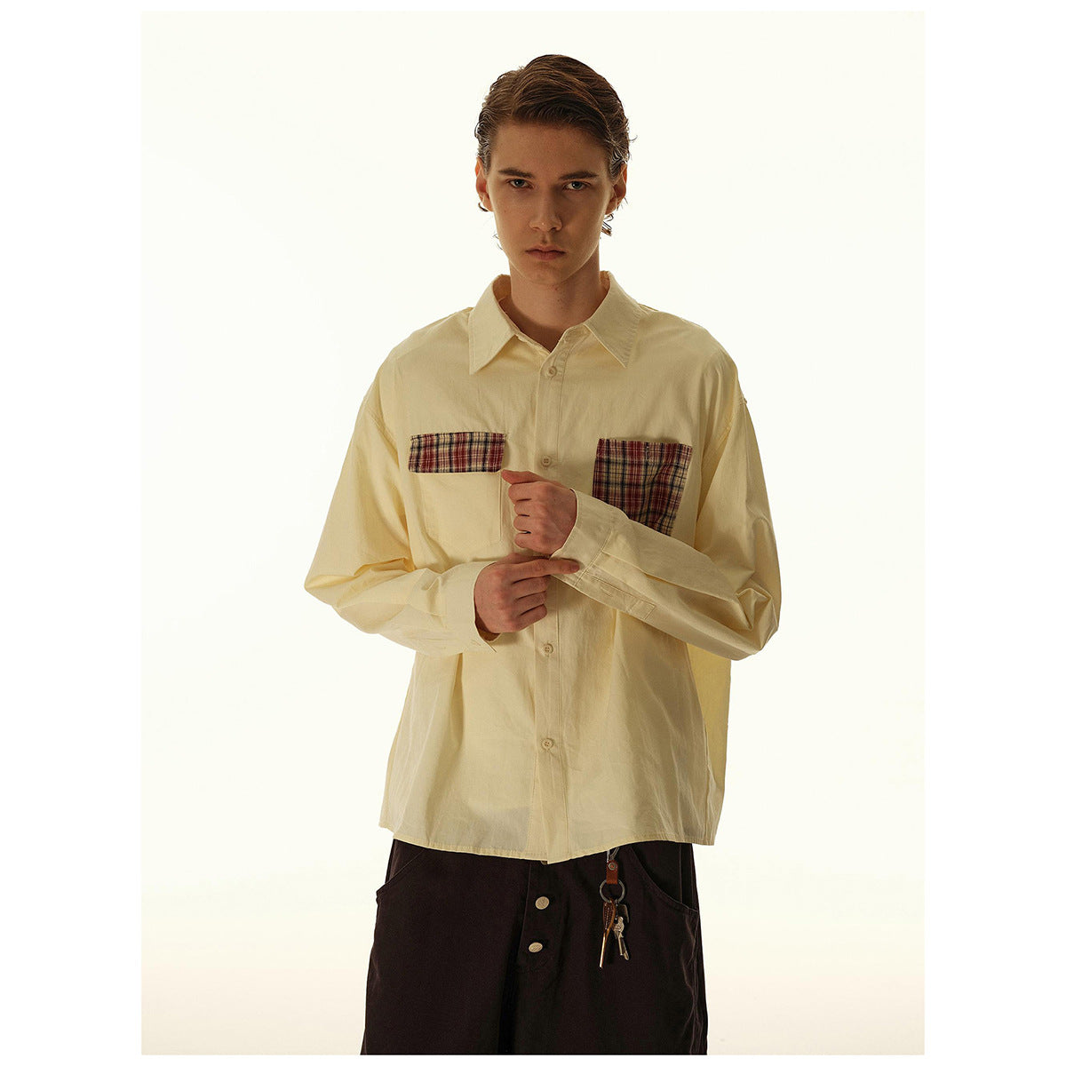 Model wearing a cream plaid-pocket shirt and wide dark trousers, adjusting one sleeve.