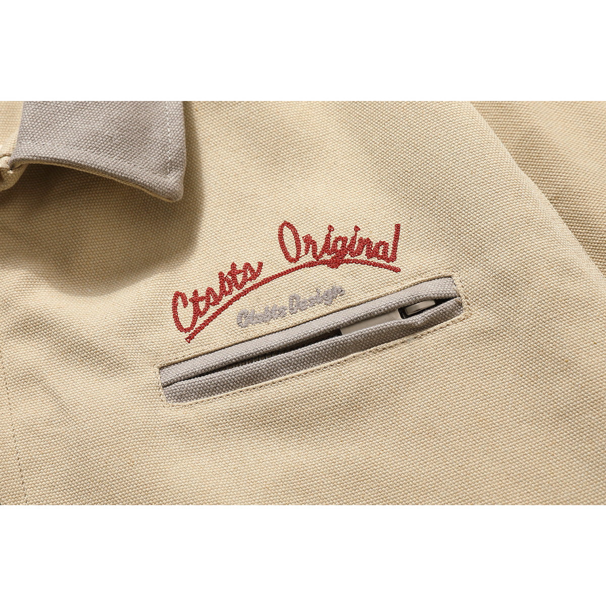 Close-up of a beige jacket's taupe-trimmed zip pocket and red embroidered script.
