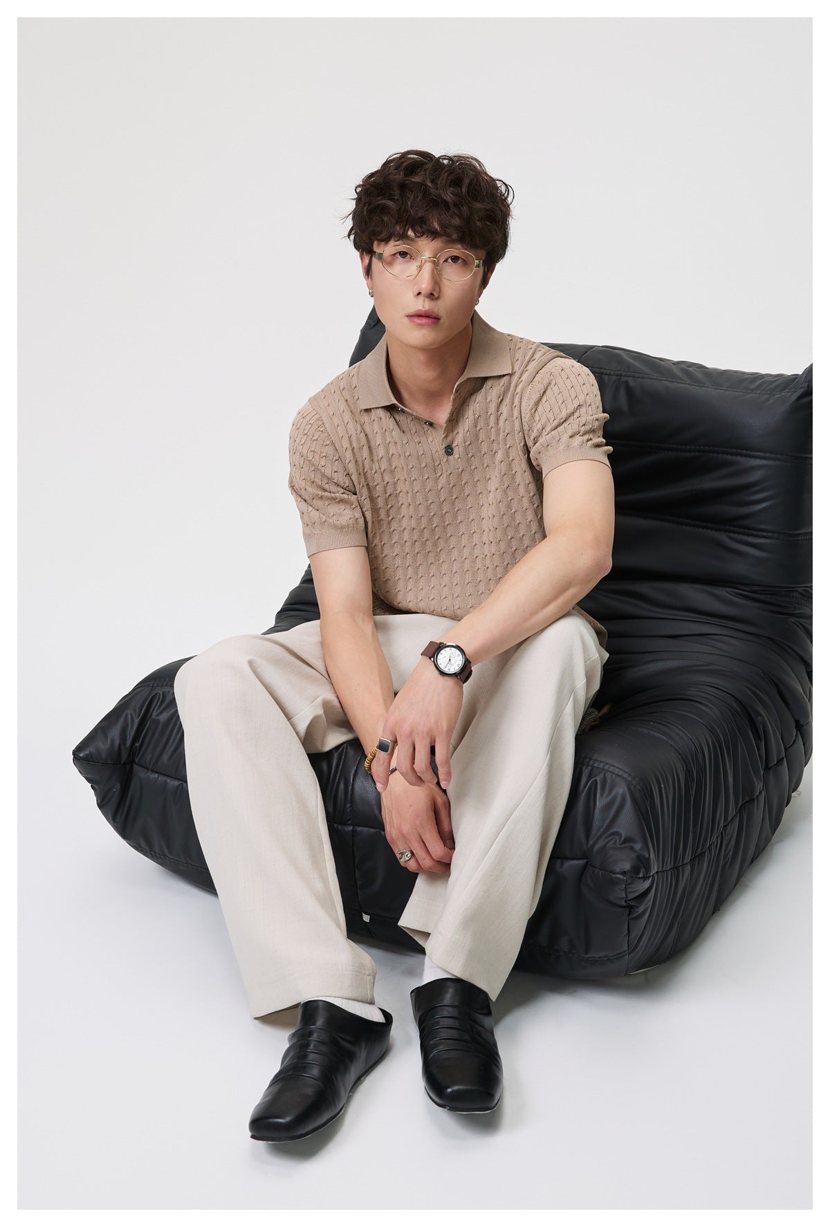 Model seated in a black chair wearing a taupe textured short-sleeve polo and light trousers.