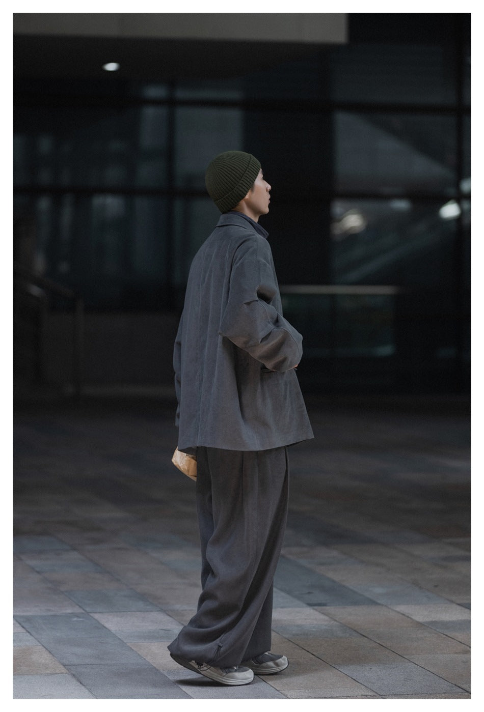 Side-back view of a man wearing an oversized charcoal-gray blazer, wide-leg trousers, and an olive ribbed beanie.