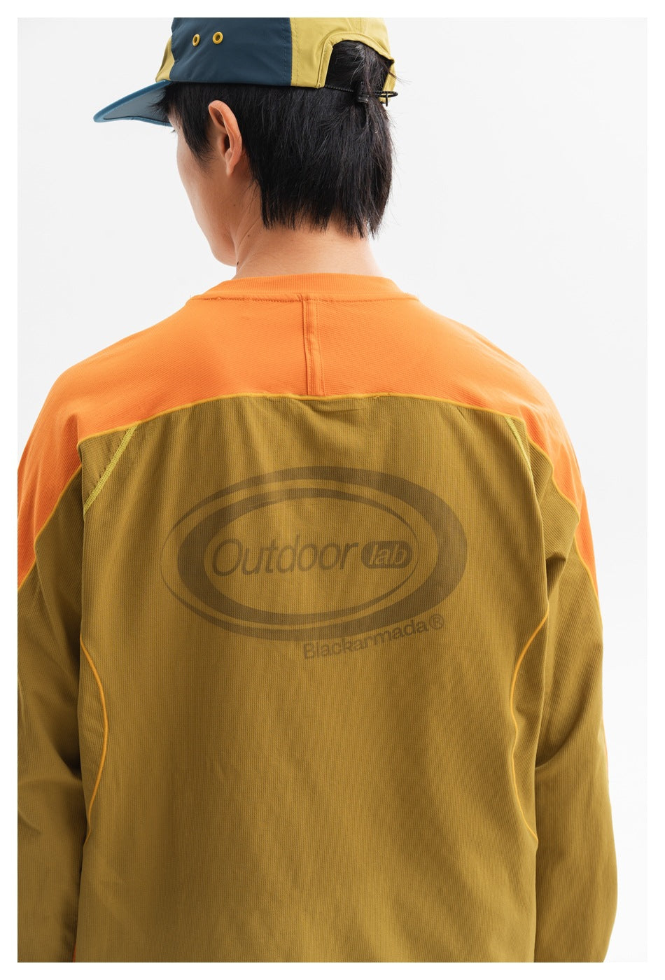 Back view of a model wearing an olive-brown top with orange shoulders and sleeves, paired with a color-block cap.