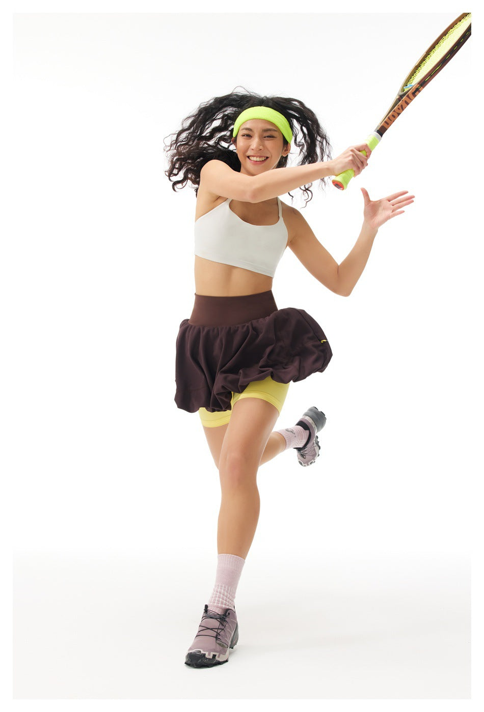 Woman swinging a racket in a dark plum tennis skirt over neon yellow shorts, white crop top and lime headband.