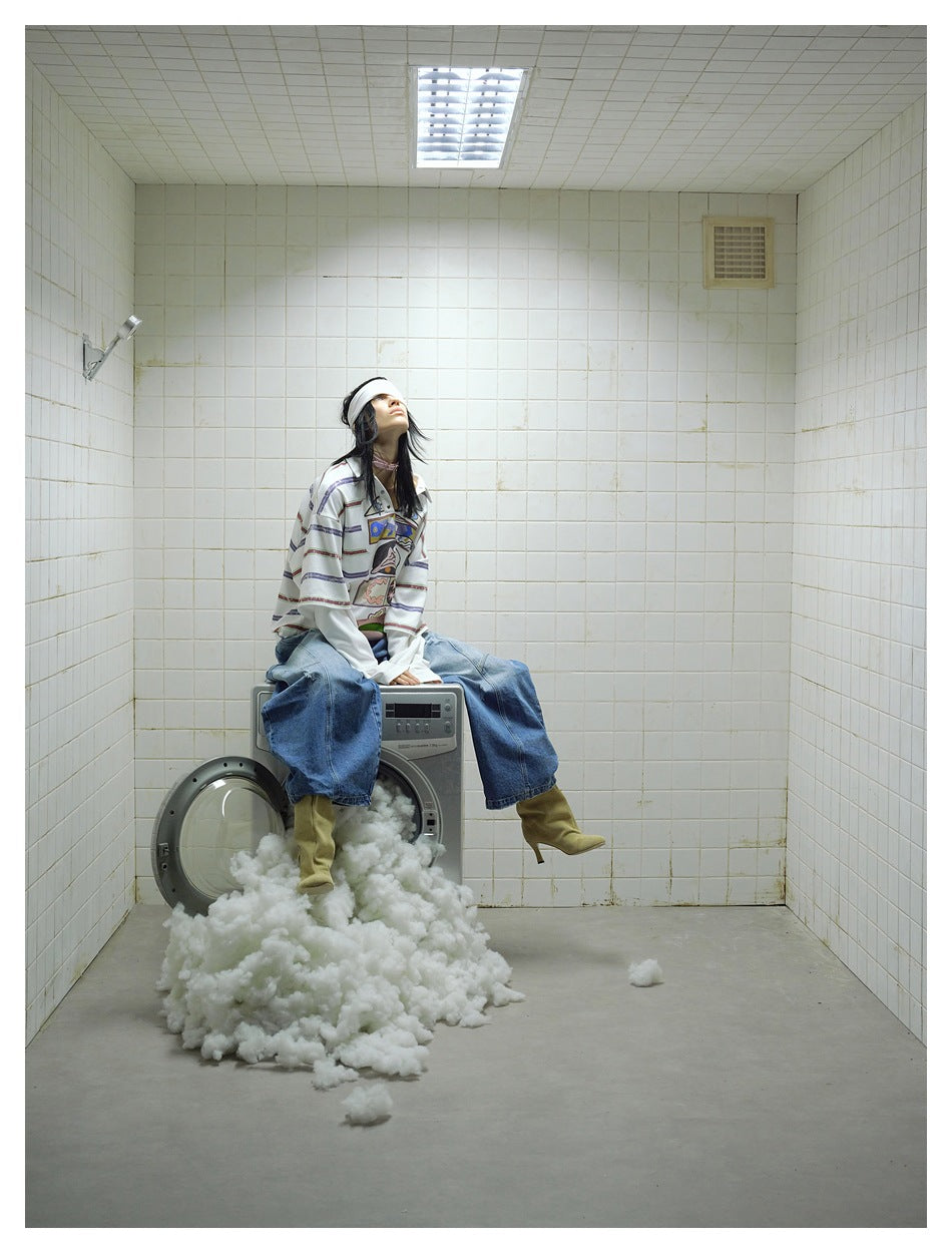 Seated model wearing a white graphic polo with long undersleeves and loose blue jeans.