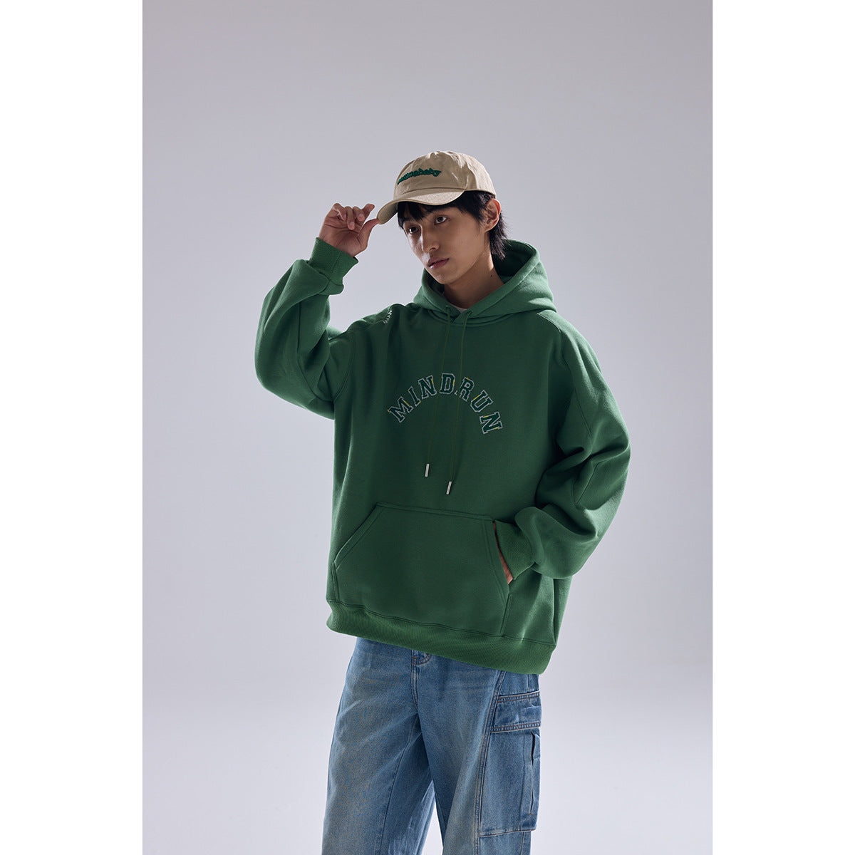 Model wearing a dark green hoodie with arched MINDRUN lettering, kangaroo pocket and drawstrings, with blue jeans and a beige