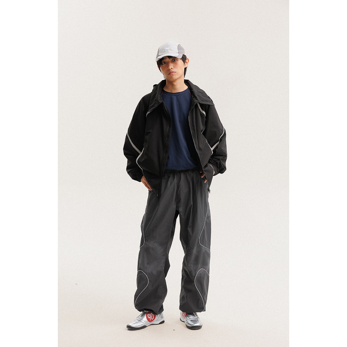 Model wearing a black jacket with white piping and loose black pants, paired with a navy shirt and white cap.