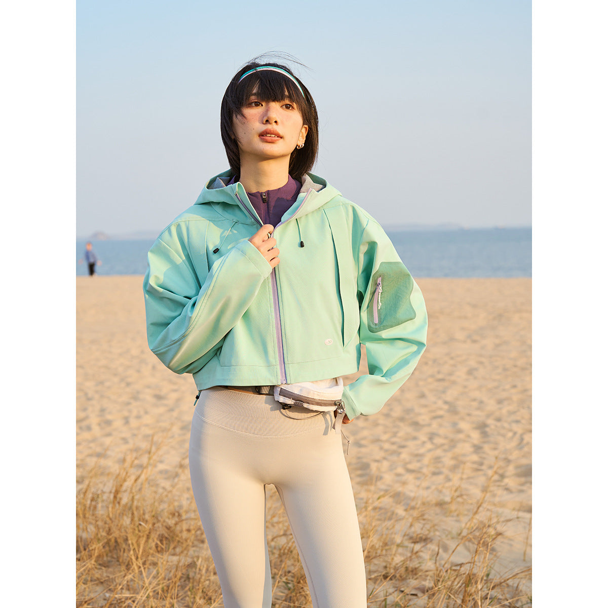 Model wearing a mint hooded cropped jacket with lavender zipper trim and a zippered sleeve pocket.