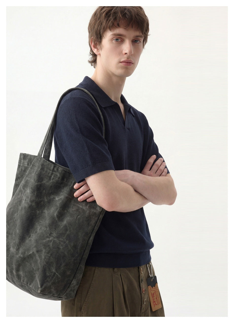 Man with crossed arms wearing a navy knit polo and carrying a large gray-green tote bag