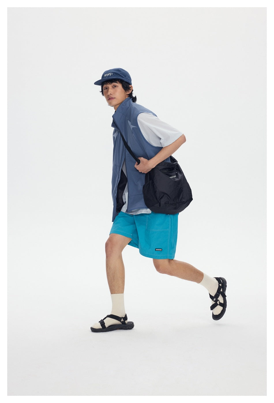 Model in a blue-gray sleeveless vest, white T-shirt, turquoise shorts, navy cap, and black crossbody bag.