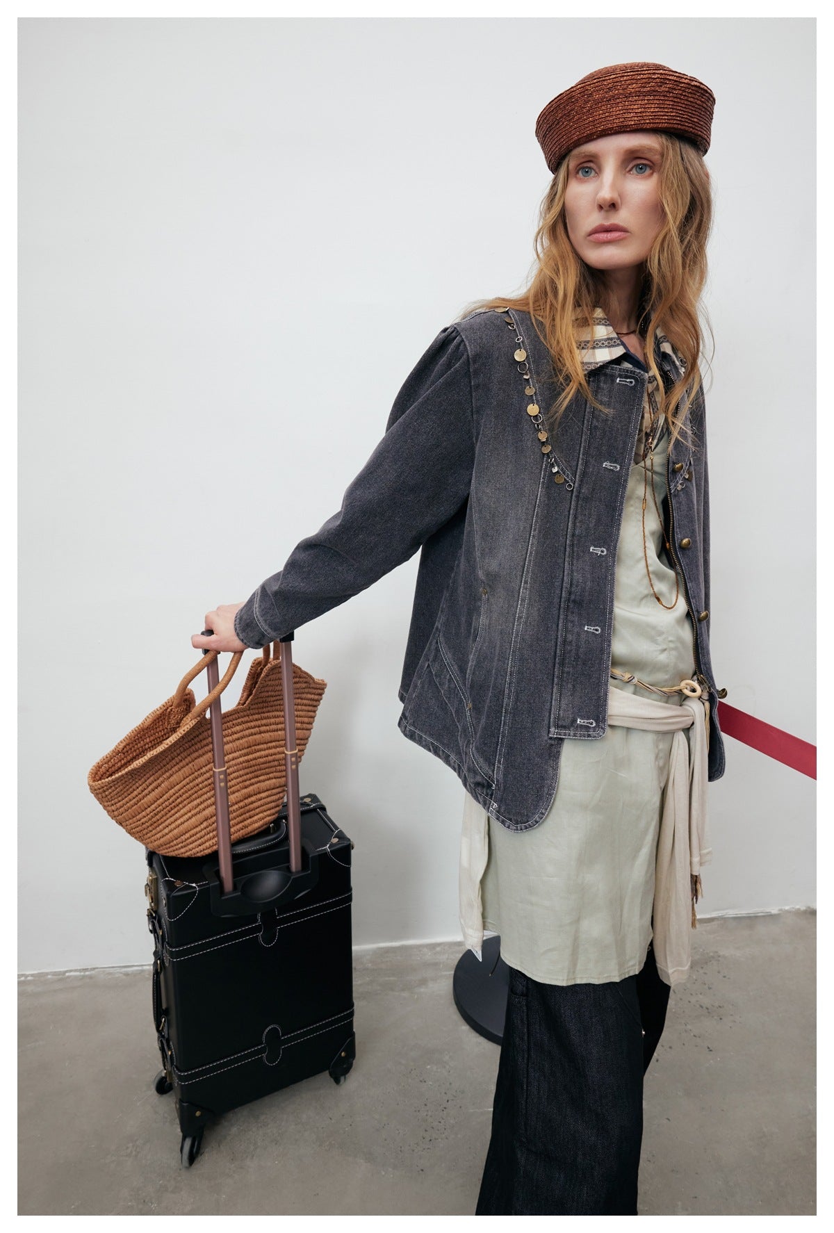 Model in a charcoal embellished denim jacket over a pale tunic, holding a woven tote beside a suitcase.