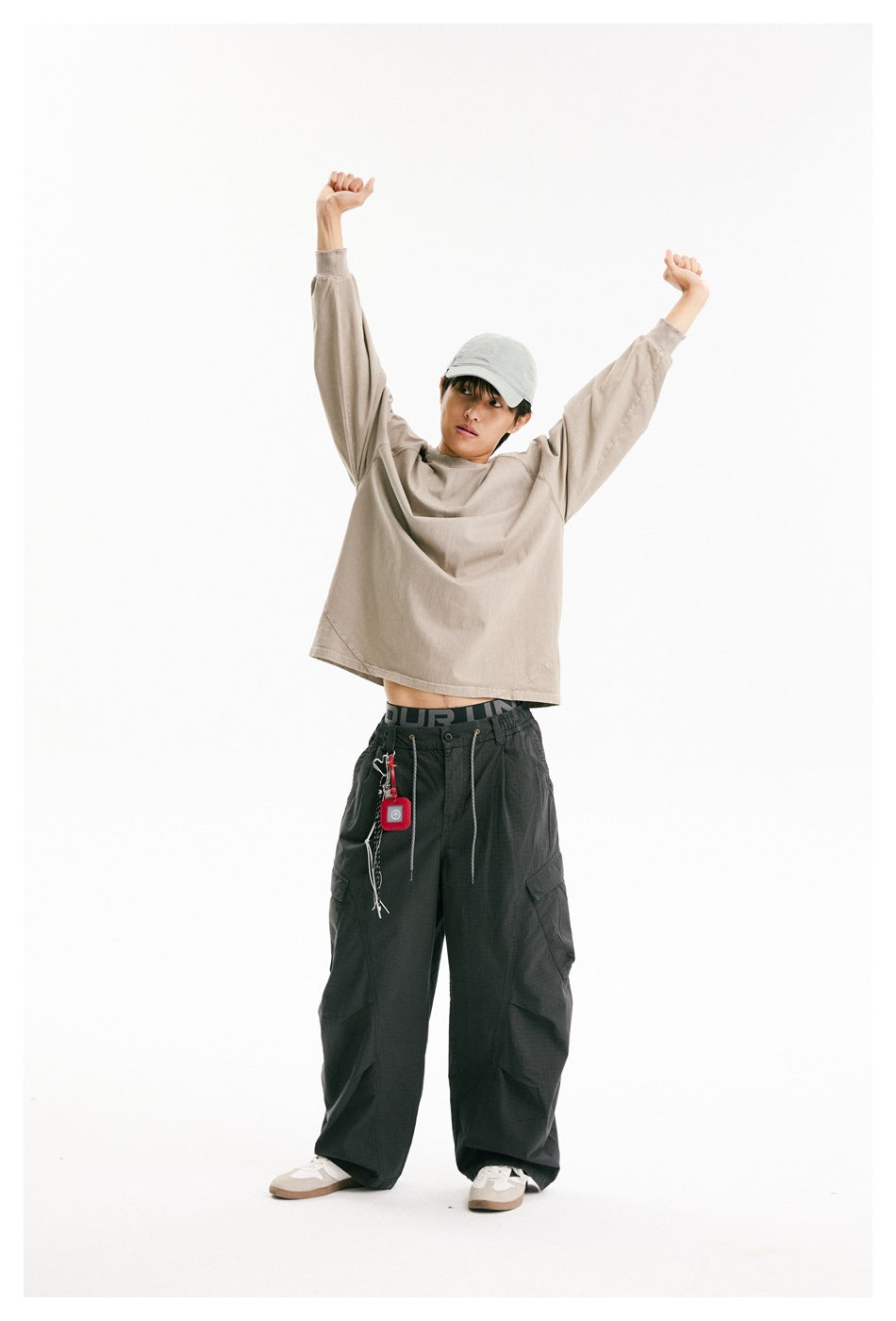 Model in an oversized taupe raglan sweatshirt and cap with dark cargo pants, standing with arms raised.