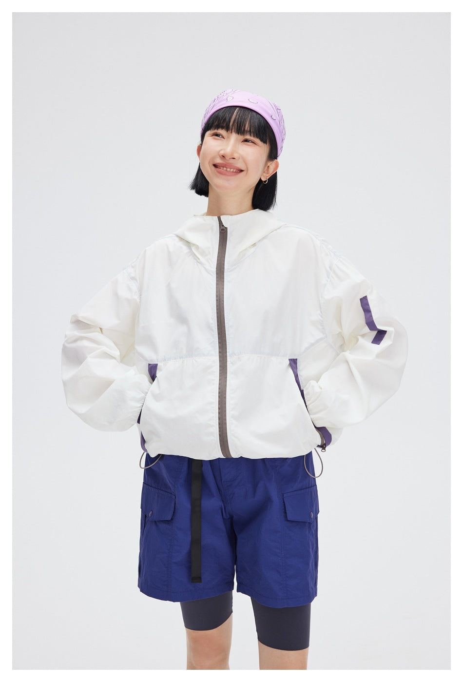 Model wearing a white hooded jacket with purple accents, paired with blue cargo shorts and dark leggings.