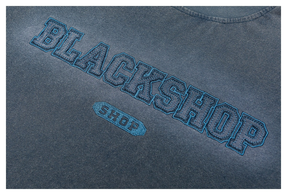 Blue-gray shirt close-up showing raised blue-outlined collegiate lettering embroidery.