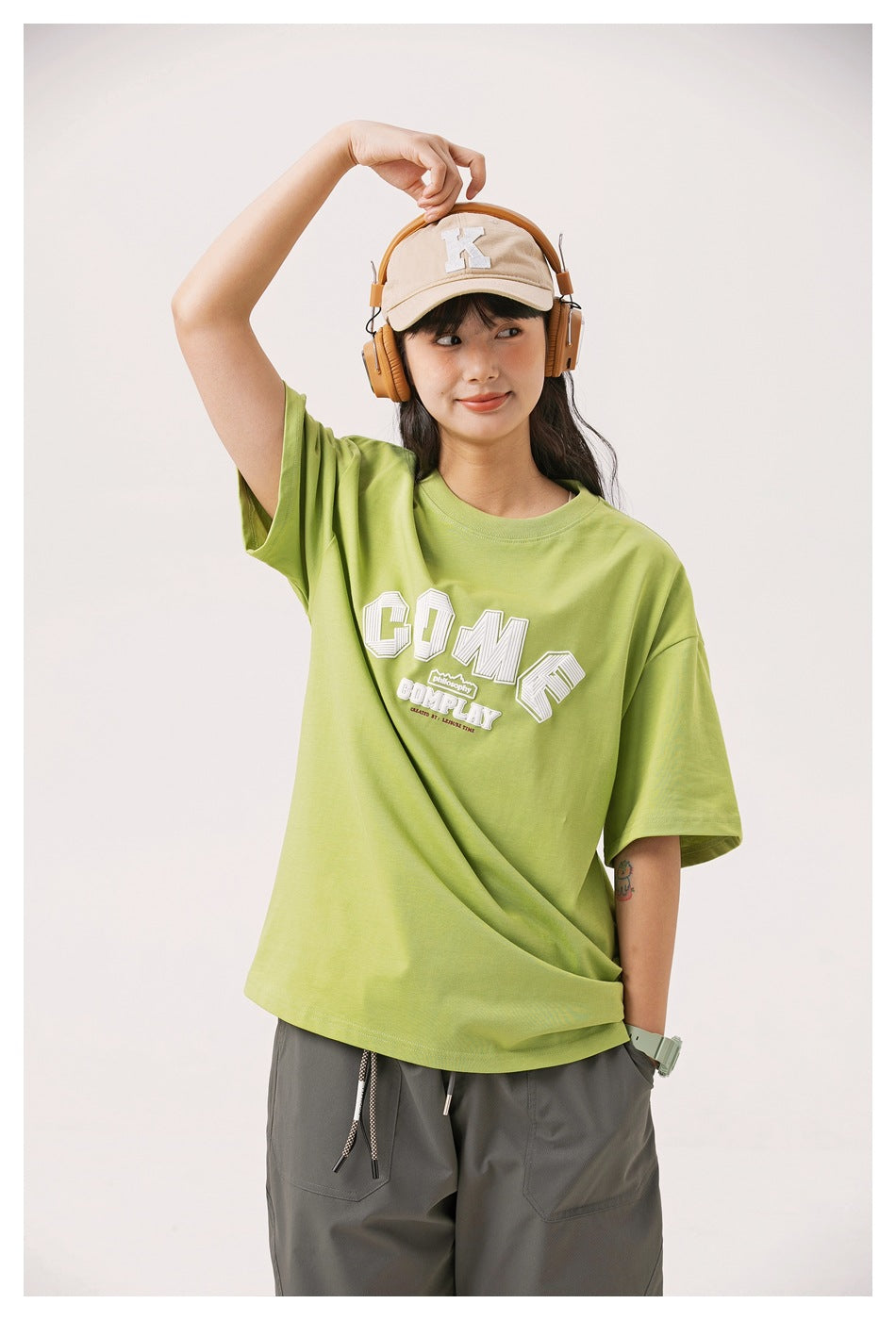Model wearing an oversized lime-green T-shirt with white collegiate-style lettering, gray pants, a beige cap, and headphones.
