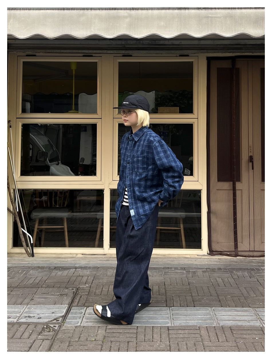 Person standing in profile wearing the washed blue plaid shirt over a striped top with wide dark jeans and a cap