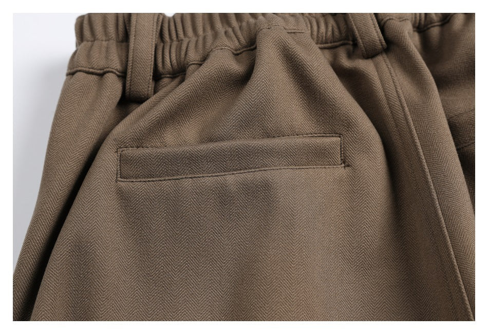 Close-up of brown trousers showing a gathered waistband, belt loops, and a stitched welt pocket.