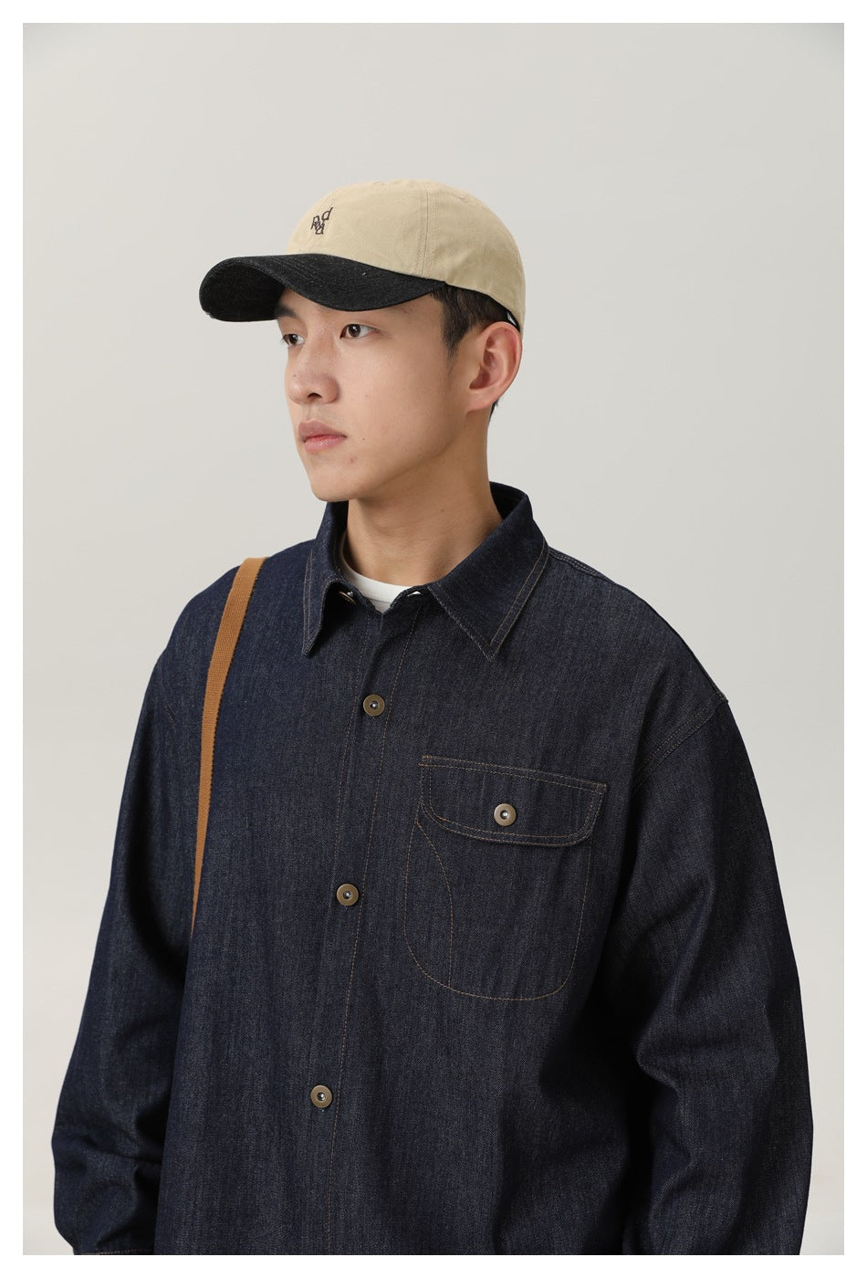 Model wearing a dark indigo denim button-up shirt with a chest pocket, paired with a beige cap with a black brim.