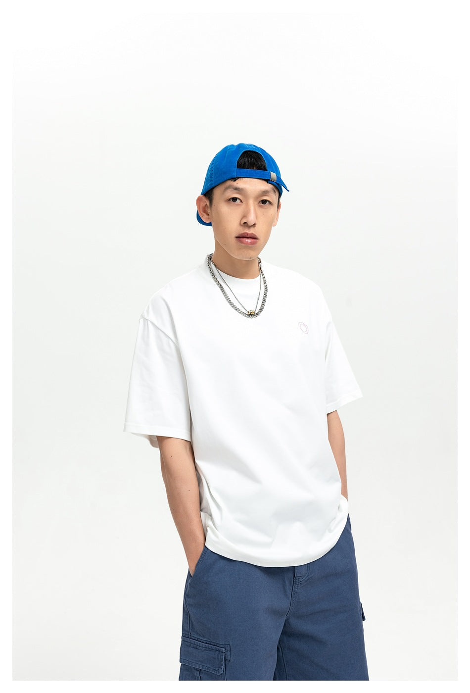 Closer front view of a man wearing a white oversized T-shirt with a small chest emblem and blue cargo pants.