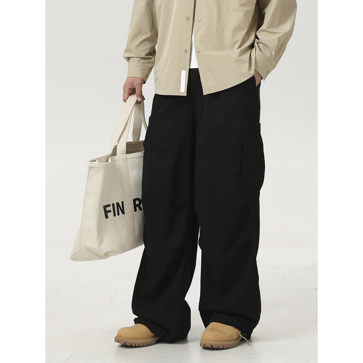 Cropped outfit view of a model in a light beige shirt and black cargo pants, carrying a printed cream tote.