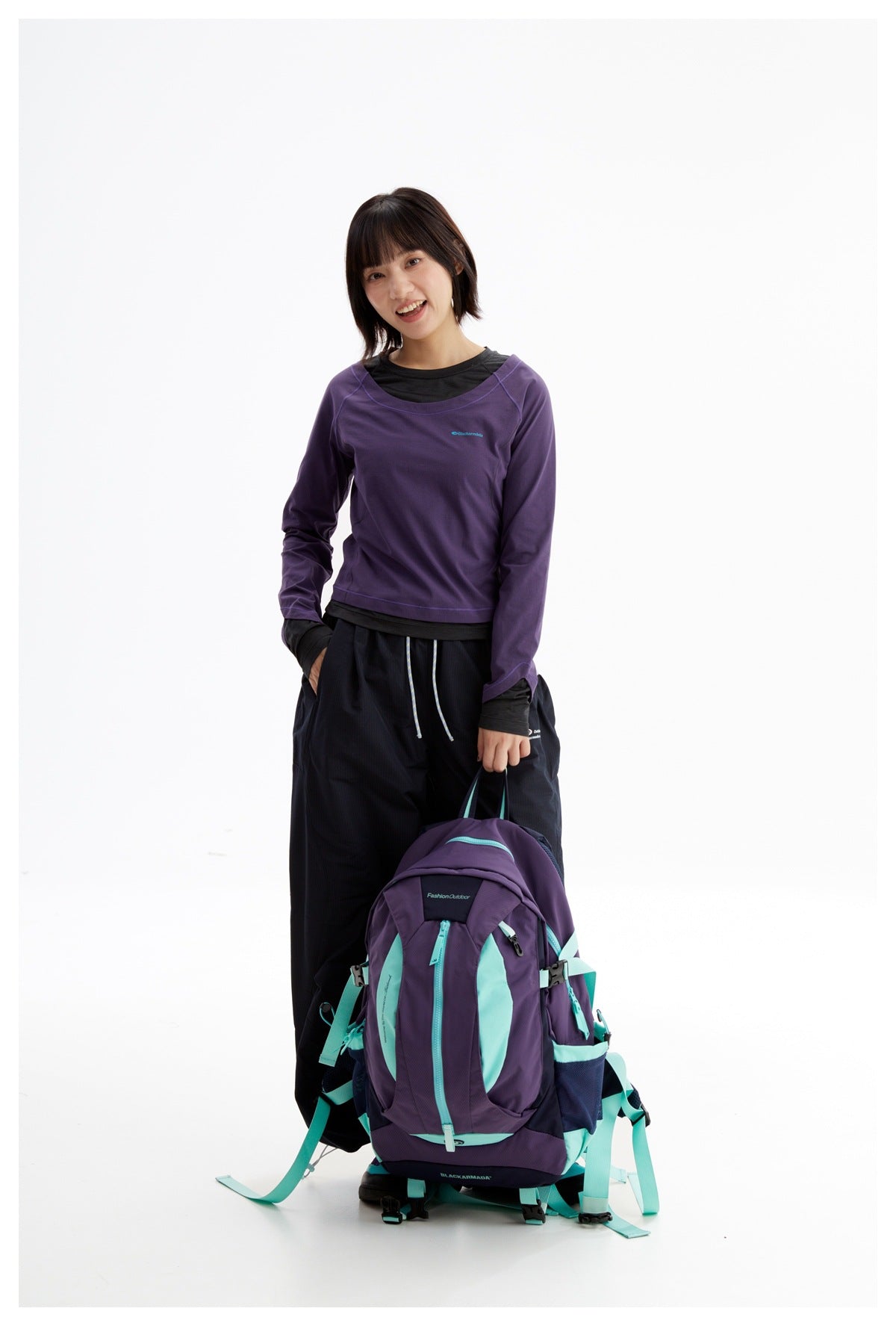Model wearing a purple long-sleeve top over a black shirt, holding a purple and turquoise hiking backpack.