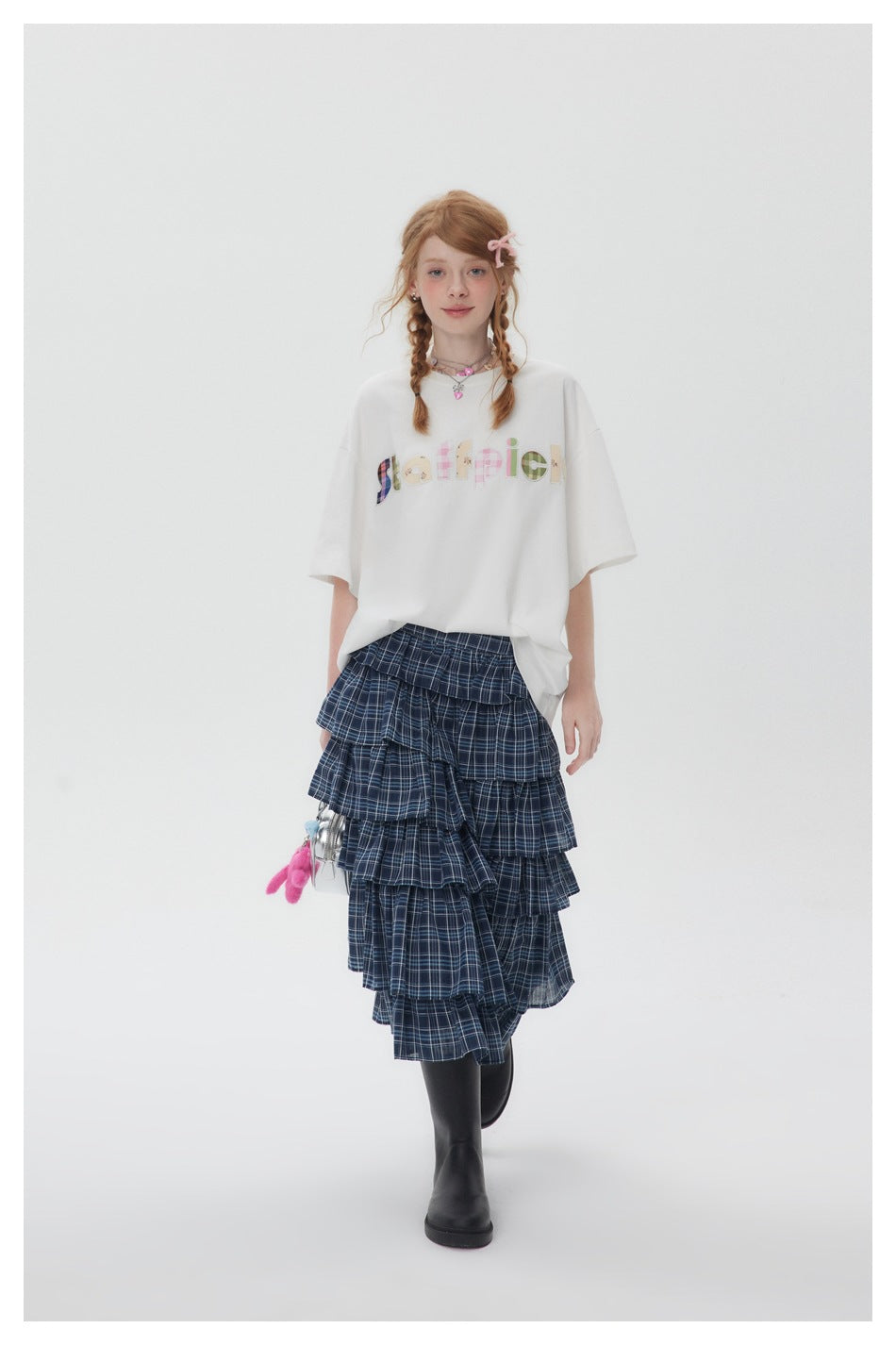 Full-length model wearing an oversized white graphic T-shirt, navy plaid tiered skirt, and black boots.