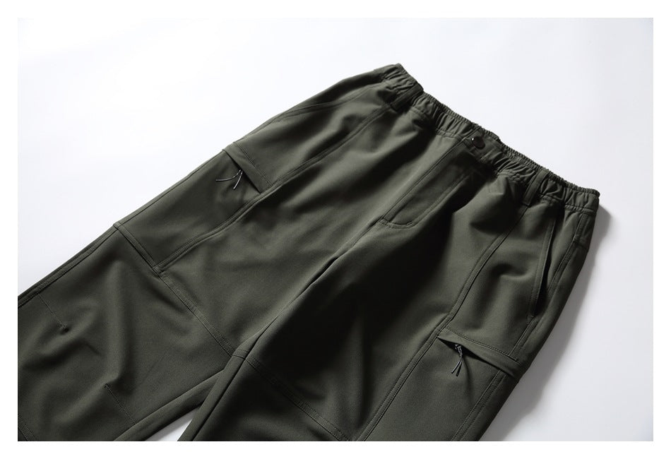 Dark olive utility pants shown from the front, with an elastic waist, button, angled pockets, zip pockets, and panel seams.