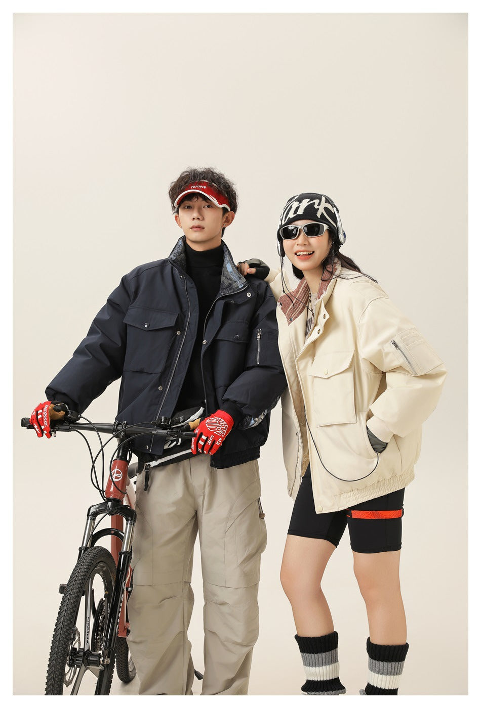 Two models pose with a bicycle, wearing navy and cream utility jackets with large flap pockets and zip details.