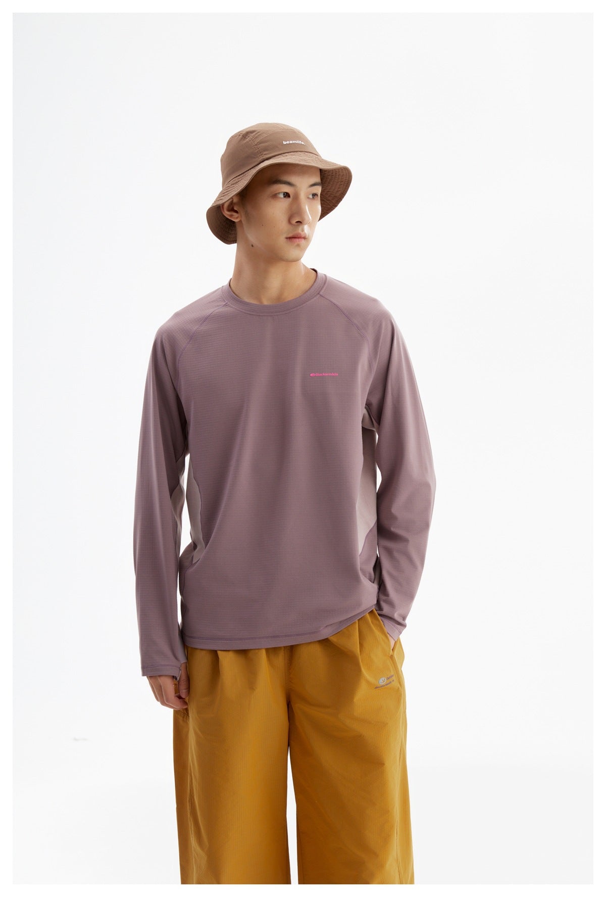 Model in a mauve long-sleeve shirt with pale side panels, mustard pants, and a brown bucket hat.