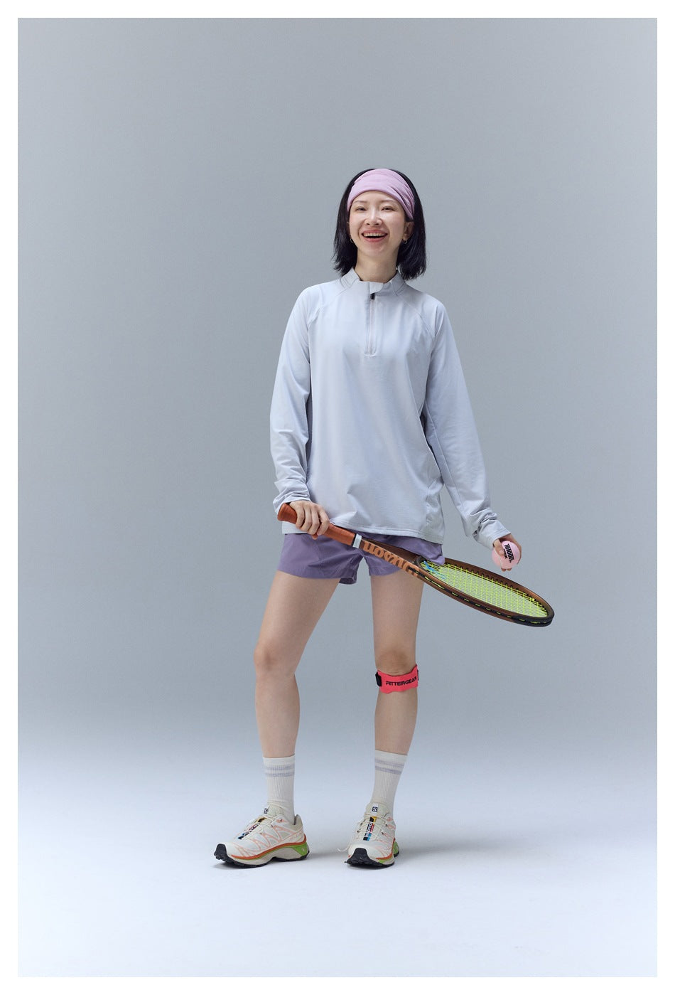Model in a pale gray quarter-zip athletic top and lavender shorts, holding a tennis racket and ball.
