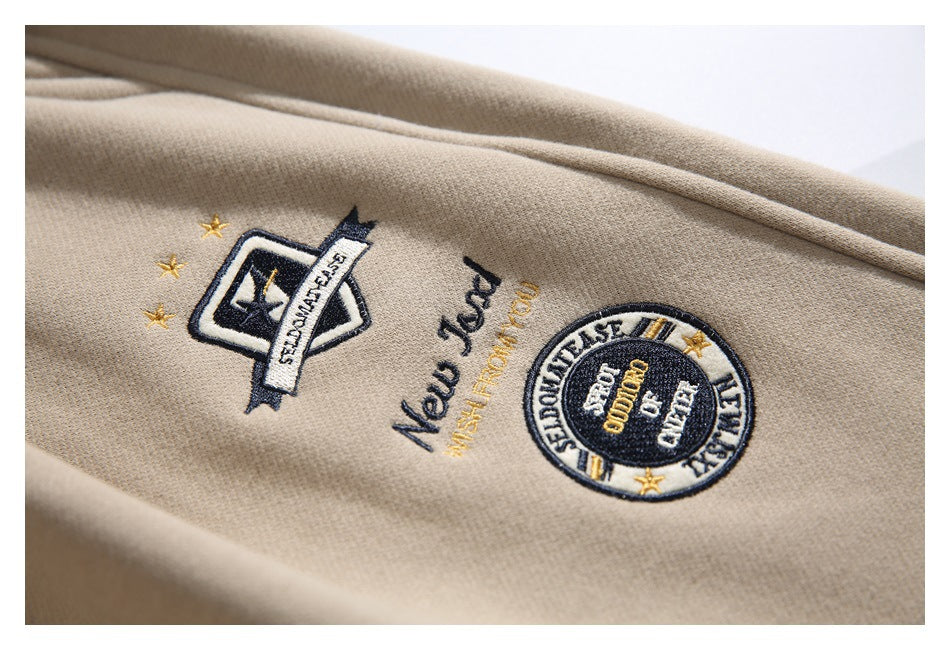 Close-up of beige jogger pants with embroidered navy, white, and gold English-text patches.