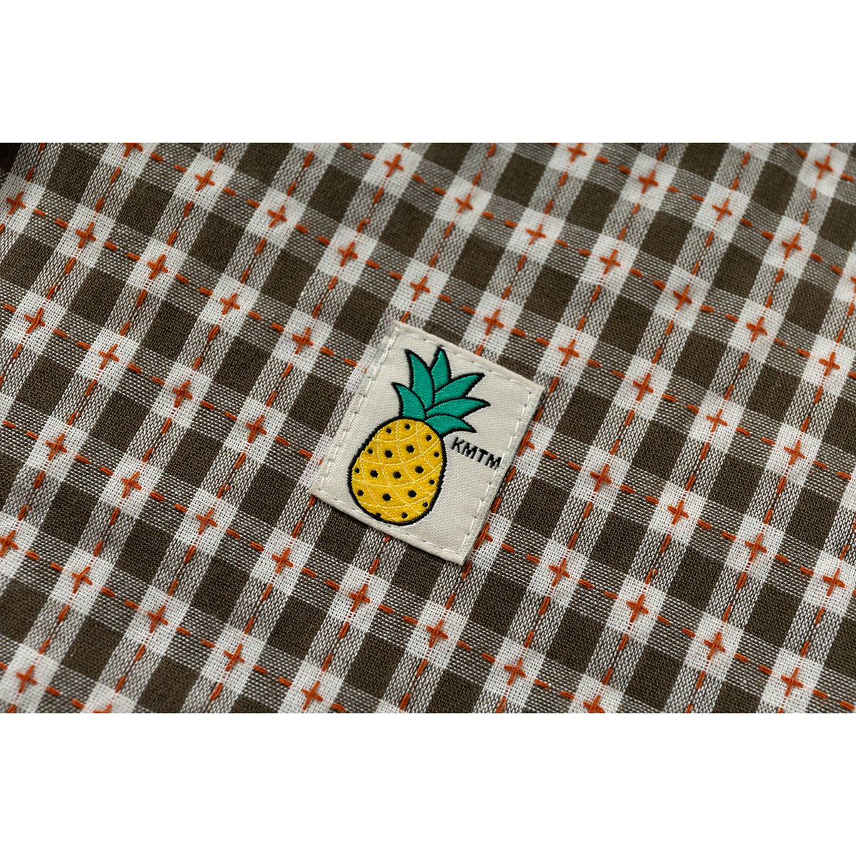 Close-up of brown and white plaid fabric with rust stitching and a small embroidered pineapple patch.