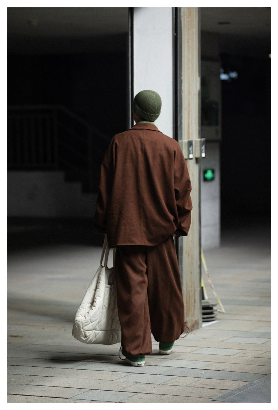 Back view of a person in a brown oversized jacket and matching trousers with a green beanie, carrying a quilted tote bag