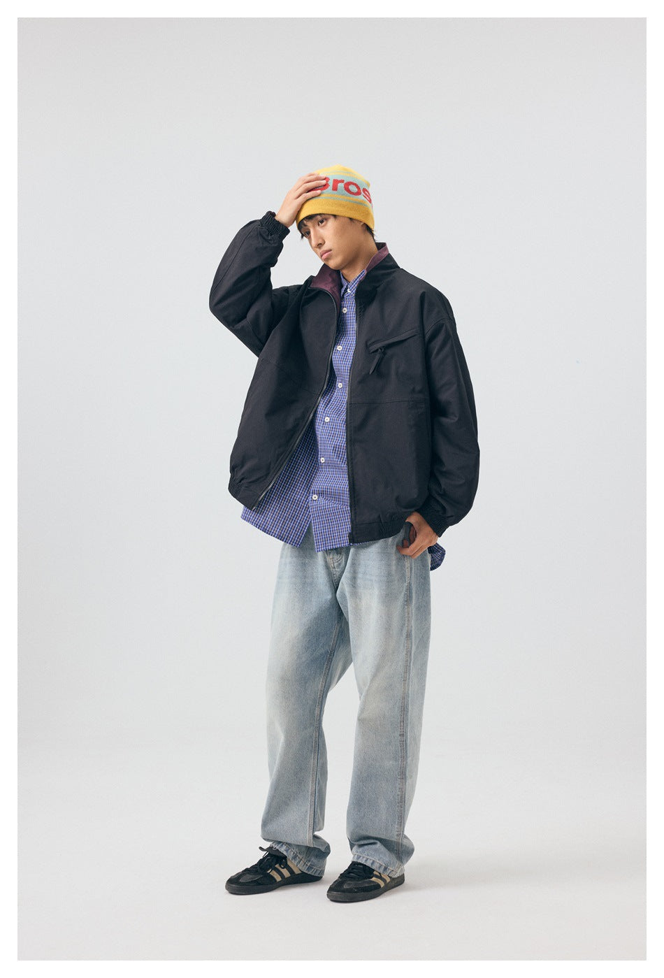 Model wearing black zip jacket over a blue plaid shirt with light wash jeans, yellow beanie and sneakers, front view